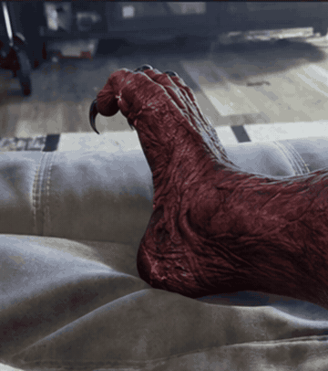 A red, sinewy demonic foot with long black claws slowly spreads and curls its toes while resting on a gray couch.